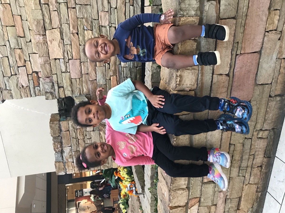 Three siblings sitting on a stone fountain ledge at the mall — Yohannes on the right grinning wide in a navy moose shirt and orange shorts