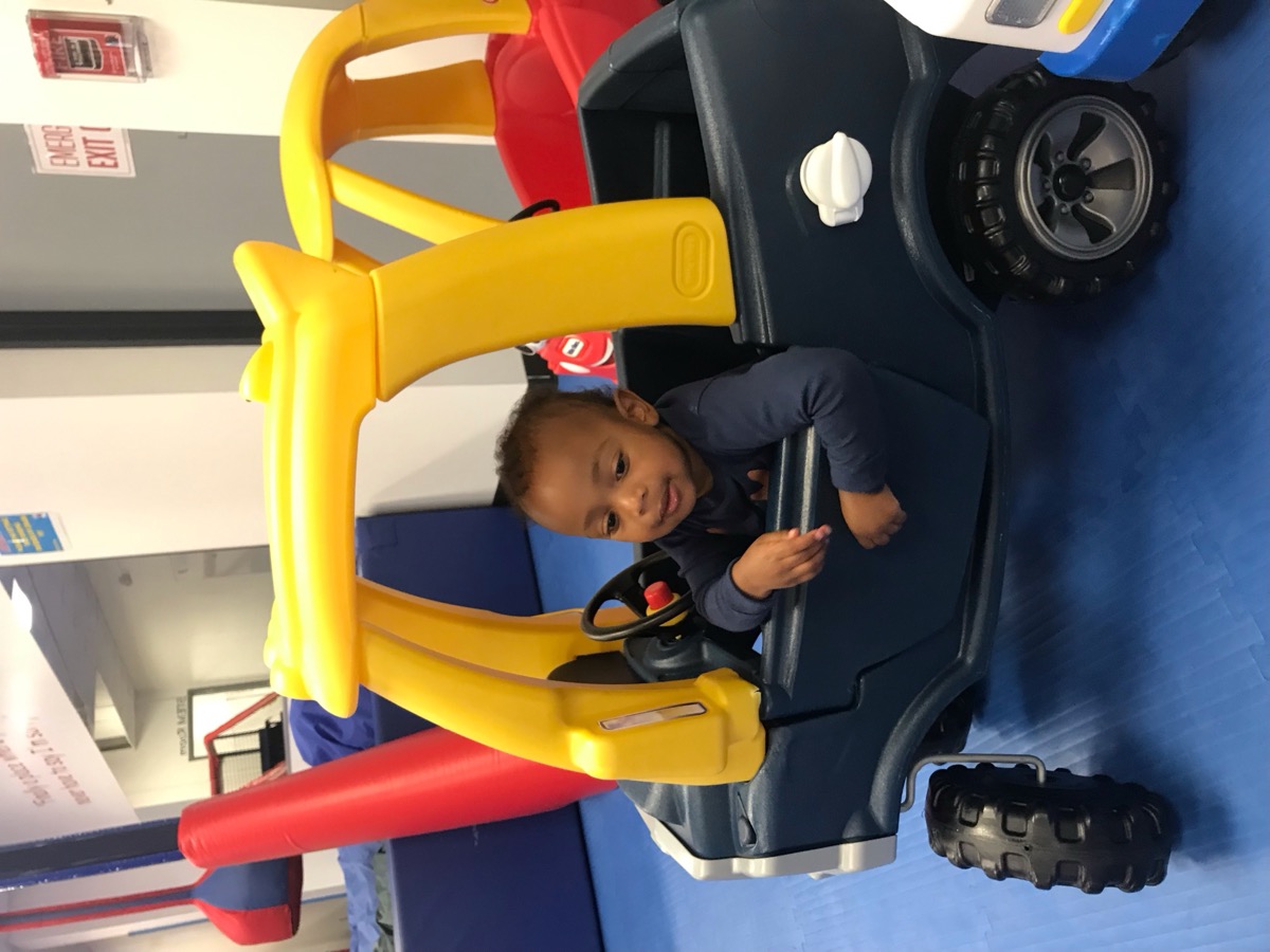 Baby Yohannes sitting inside a navy and yellow Little Tikes toy truck at an indoor play area