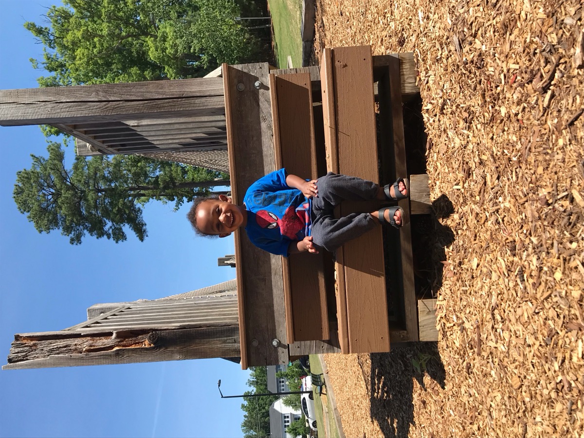 Yohannes sitting on a wooden bench at a playground, beaming in a blue Spider-Man t-shirt on a sunny day