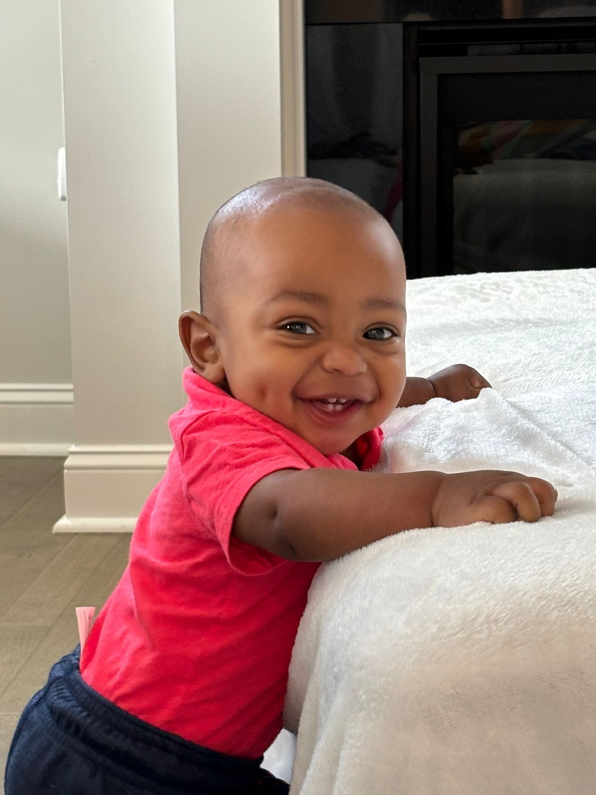 Baby Yohannes in a coral red polo pulling himself up on a white blanket, flashing a big happy grin