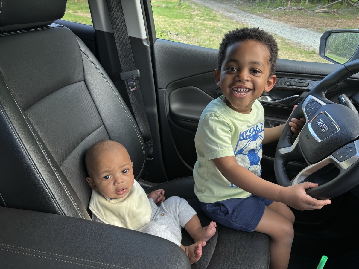 Baby Yohannes and his older brother sitting together in the front seat of a car, brother holding the steering wheel and both smiling
