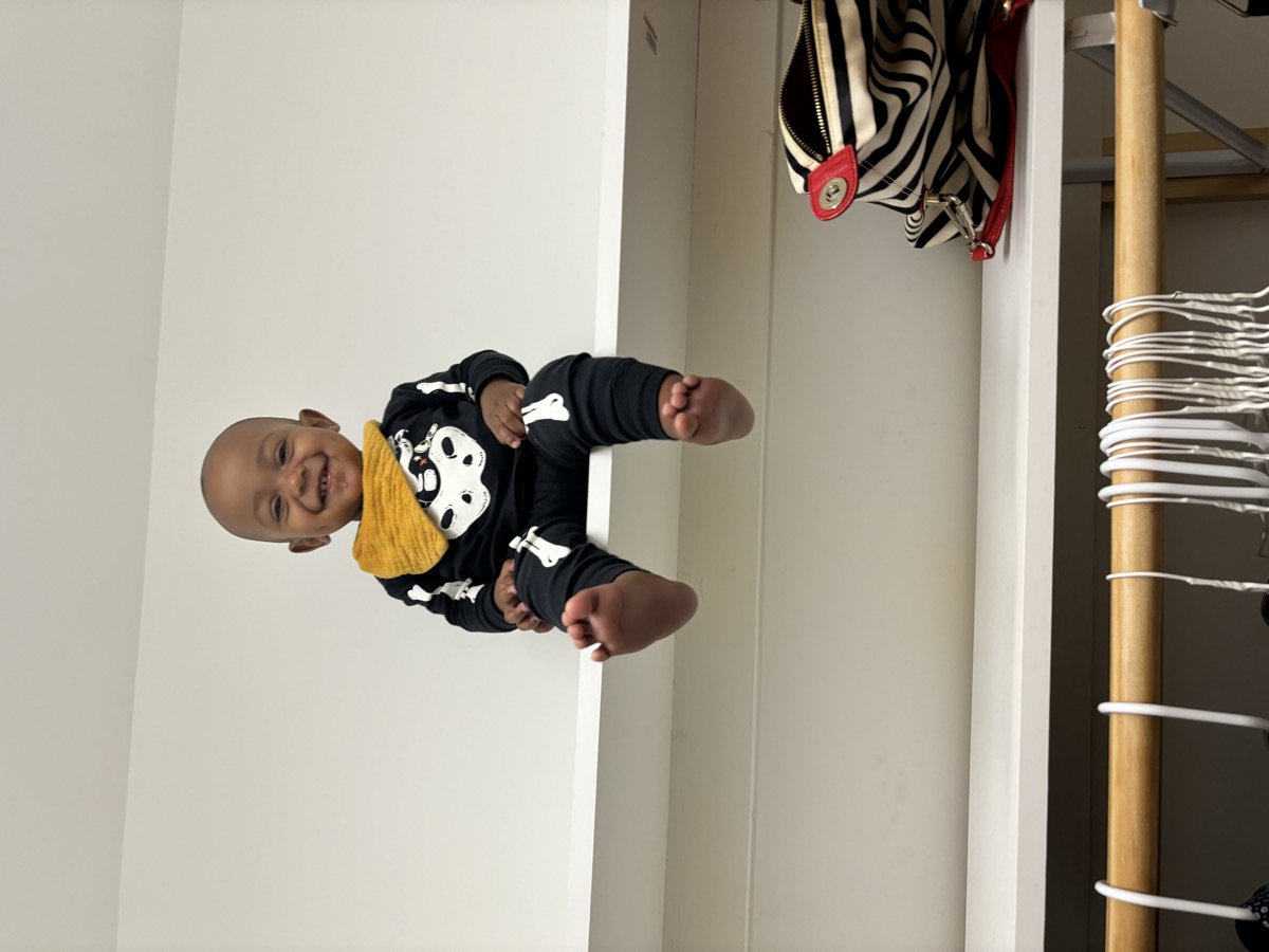 Baby Yohannes sitting high up on a white closet shelf in a black skeleton outfit with a mustard bandana bib, grinning mischievously