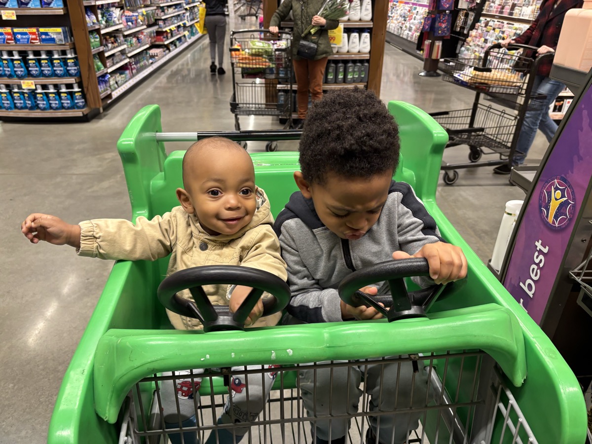 Baby Yohannes and his older brother riding together in a green race-car grocery cart at the store