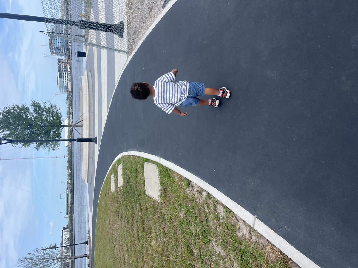 Toddler Yohannes walking alone on a waterfront path in a striped shirt and denim shorts, seen from behind
