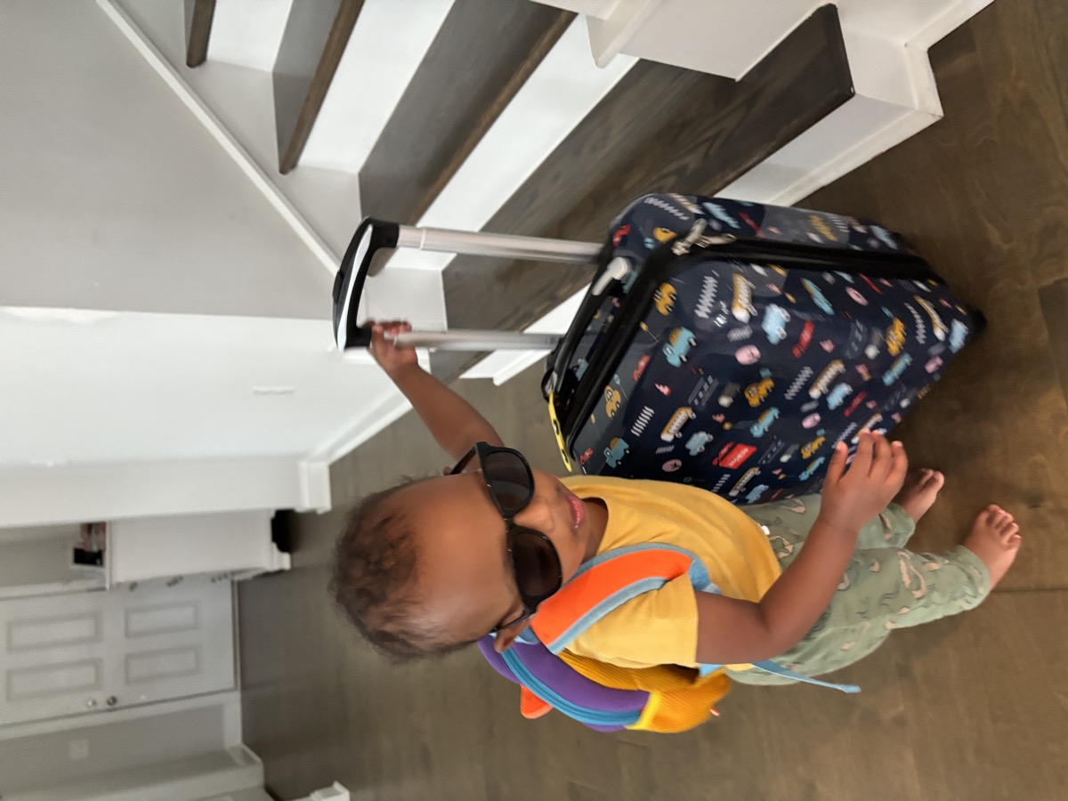 Toddler Yohannes wearing dark sunglasses and a colorful backpack, pulling a vehicle-print suitcase by the stairs — ready to travel