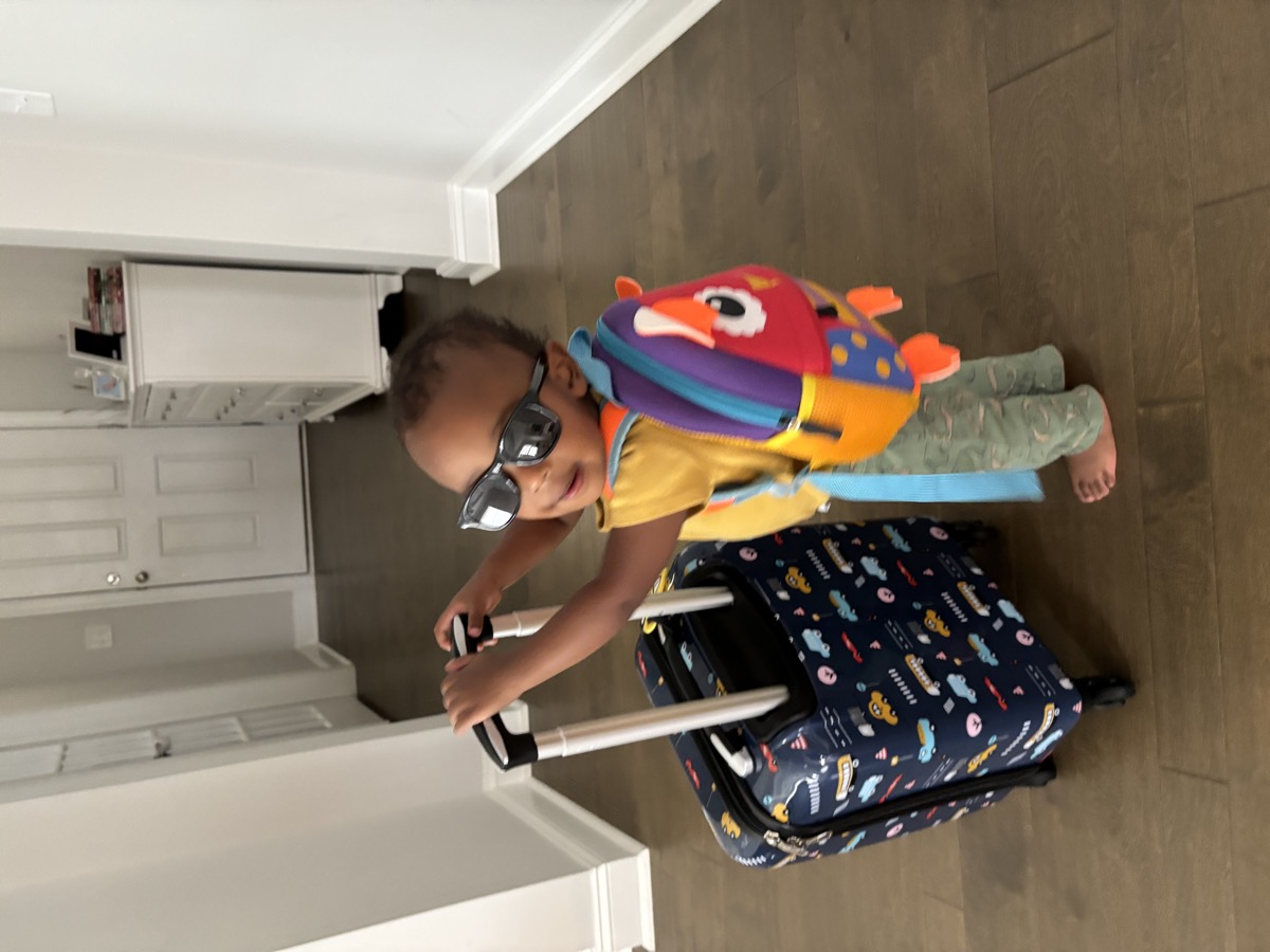 Yohannes in sunglasses and a yellow shirt holding onto his vehicle-print rolling suitcase with a colorful animal backpack — travel-ready, another angle