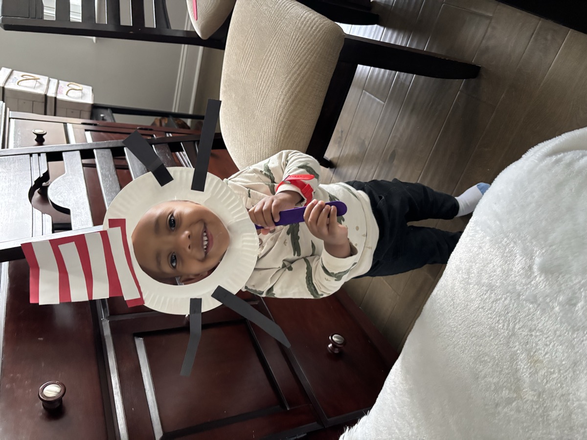 Yohannes holding a paper-plate Cat in the Hat craft up to his face, beaming in the dining room