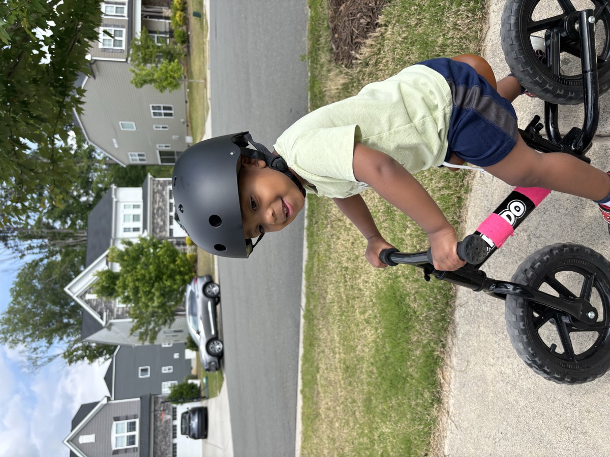 Yohannes riding a black balance bike with a helmet on, smiling on the neighborhood sidewalk