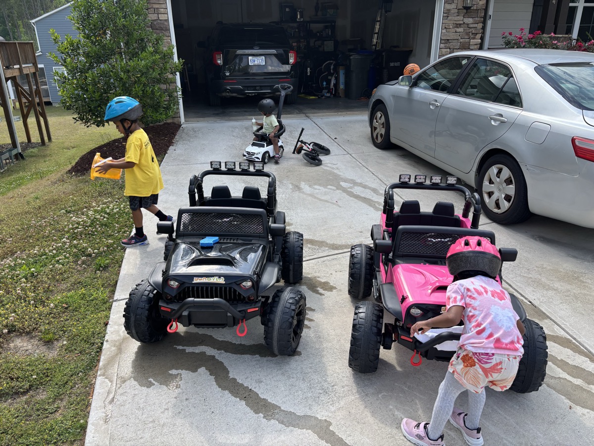 All three kids in the driveway with their ride-on power wheels — a black Jeep, a pink Jeep, and Yohannes on a white ride-on in the background