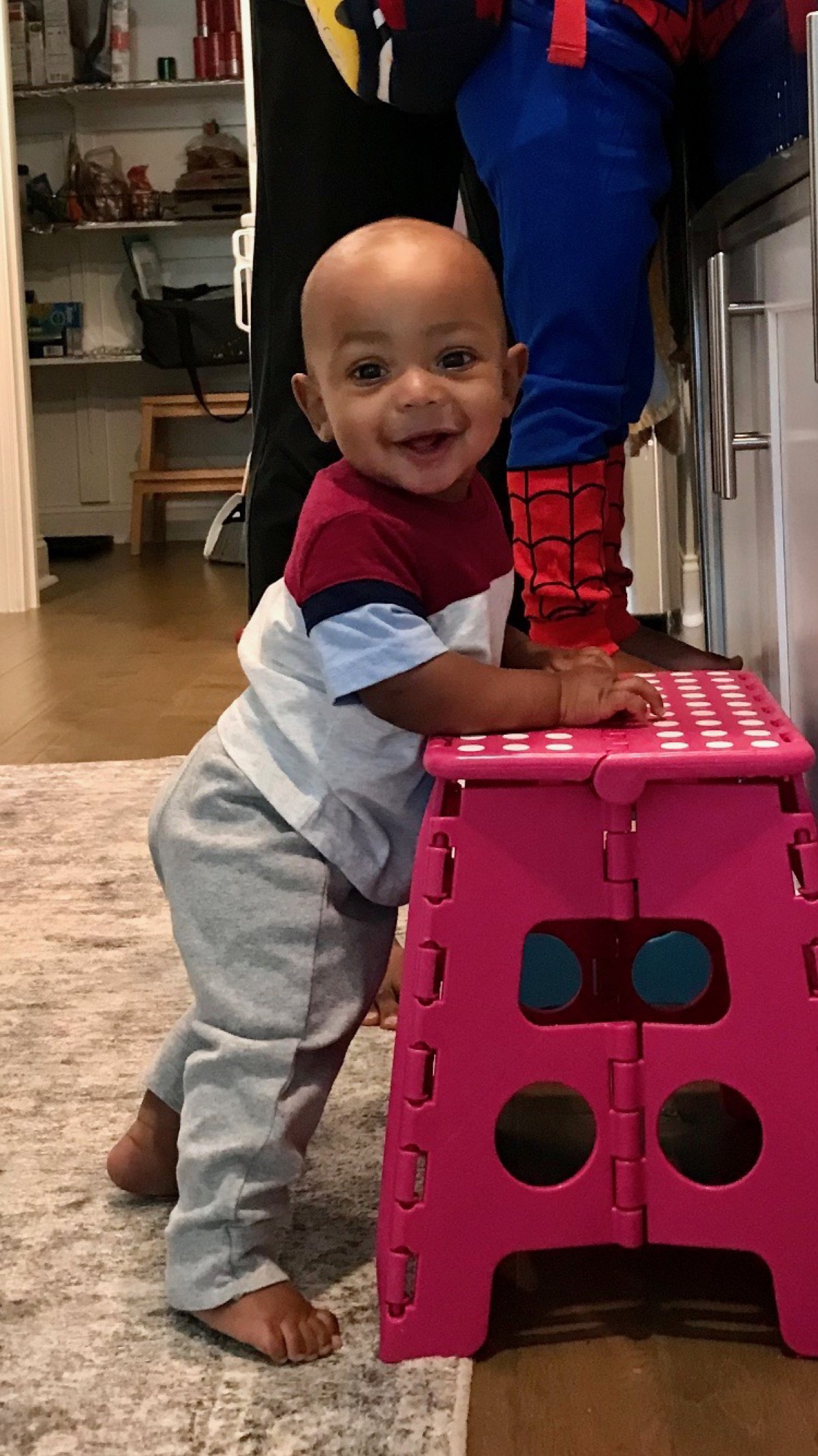 Baby Yohannes standing and leaning on a pink folding step stool in the kitchen, smiling with his brother in a Spider-Man costume behind him
