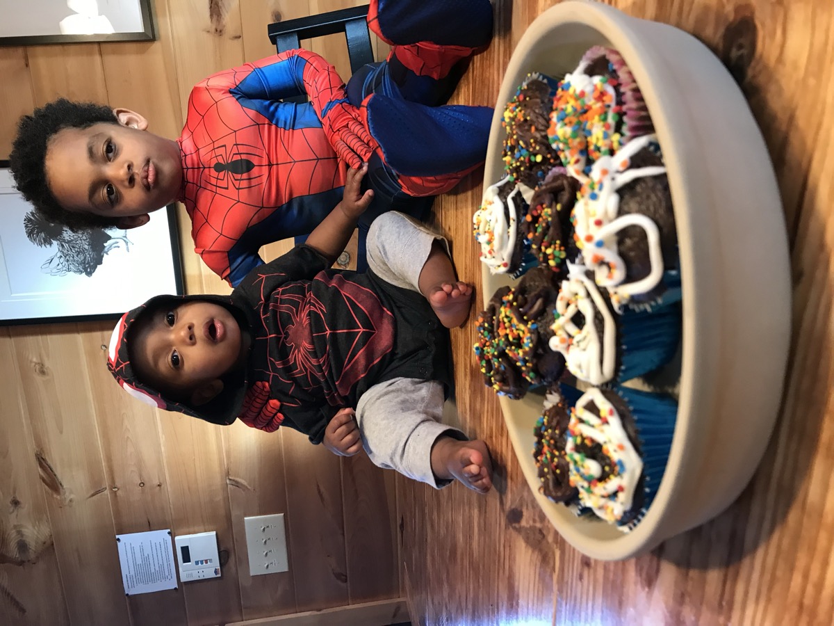 Baby Yohannes in a Miles Morales Spider-Man hoodie sitting next to his brother in a Spider-Man costume, a tray of sprinkled cupcakes in front of them