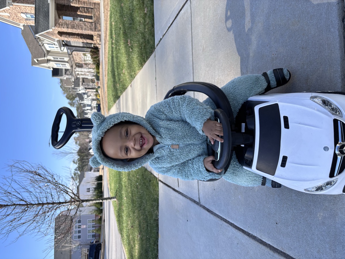 Baby Yohannes in a cozy blue bear-ear fleece onesie, grinning in a white Mercedes push car on the neighborhood sidewalk