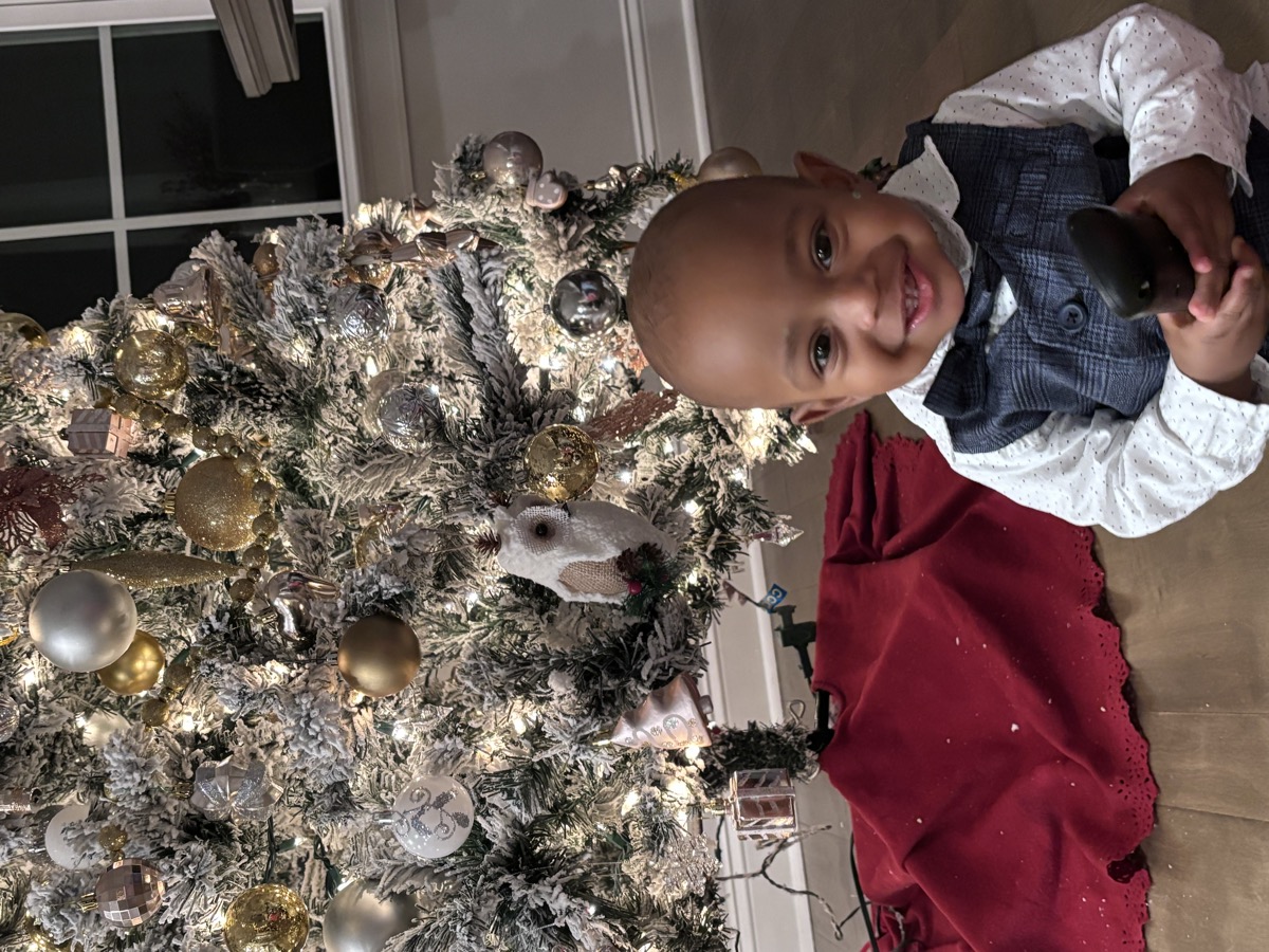Baby Yohannes in his vest and bow tie outfit smiling in front of a lit flocked Christmas tree with gold and silver ornaments