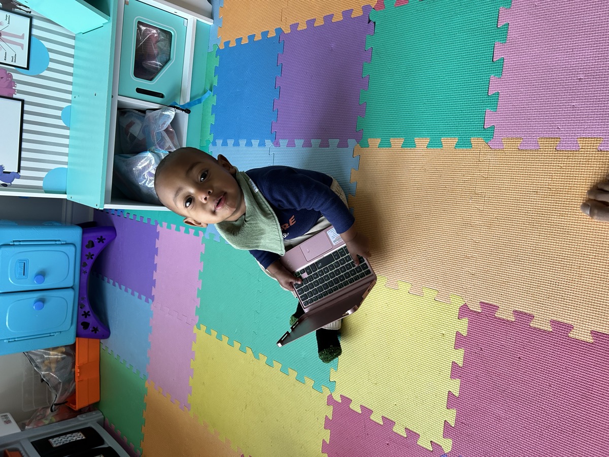 Baby Yohannes sitting on a colorful foam play mat holding a small laptop toy, looking up at the camera in a playroom