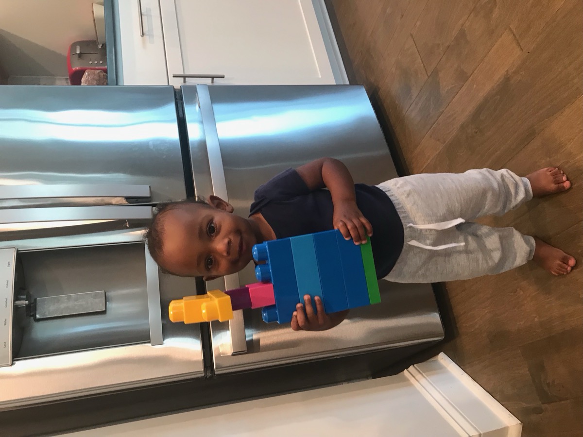 Toddler Yohannes standing in the kitchen proudly holding up a tall colorful Duplo block tower he built