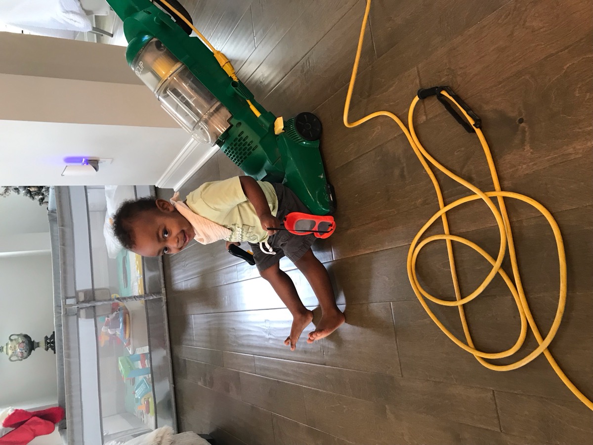 Baby Yohannes sitting on the floor grinning next to a green upright vacuum cleaner, pretending to help clean