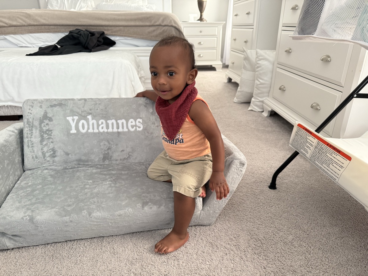 Yohannes standing next to his personalized gray couch showing the Yohannes embroidery, wearing an orange tank and burgundy bib