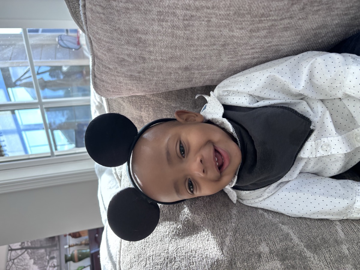 Baby Yohannes wearing Mickey Mouse ears headband and a polka-dot shirt, laughing on the couch with a black bandana bib